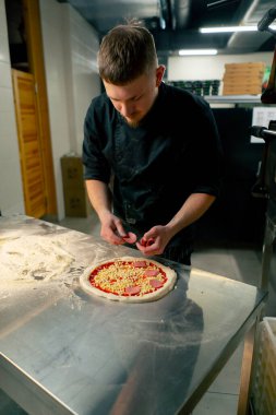 Profesyonel bir mutfakta bir pizzacıda siyah üniformalı genç bir şef pizzaya malzeme koyar.