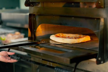 Profesyonel bir mutfakta bir pizzacıda siyahlı genç bir şef spatulayla fırın için çiğ pizza alır.