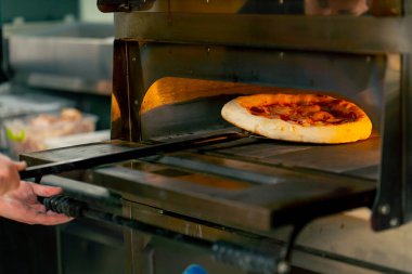 Profesyonel bir mutfakta bir pizzacıda siyahlı genç bir şef spatulayla fırın için çiğ pizza alır.