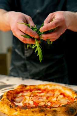 Bir pizzacının profesyonel mutfağında siyah üniformalı genç bir şef pizzanın içine malzeme koyar.