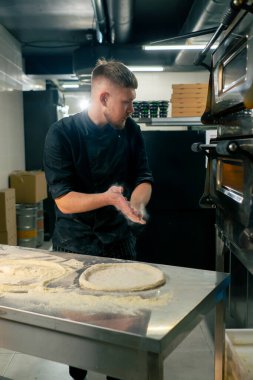 Profesyonel bir mutfakta, pizzacıda siyah üniformalı genç bir şef pizza hamuru hazırlıyor.