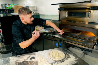 Profesyonel bir mutfakta bir pizzacıda siyah üniformalı genç bir şef fırına pizza koyar.