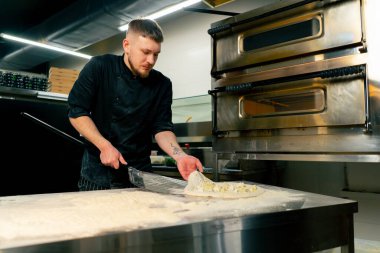 Profesyonel bir mutfakta bir pizzacıda siyah üniformalı genç bir şef fırına pizza koyar.