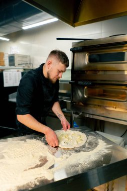 Profesyonel bir mutfakta bir pizzacıda siyah üniformalı genç bir şef fırına pizza koyar.