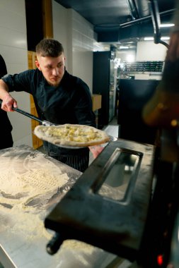 Profesyonel bir mutfakta bir pizzacıda siyah üniformalı genç bir şef fırına pizza koyar.
