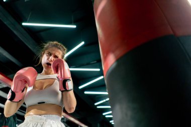 Dövüş kulübünde pembe eldivenli beyaz üniformalı genç bir kız kırmızı bir çanta üzerinde teknik tekme ve yumruk pratiği yapıyor.
