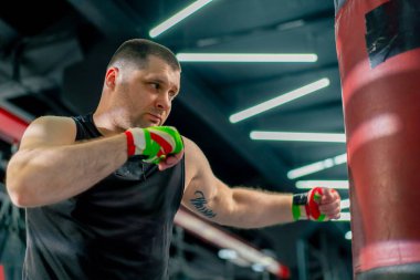 Bir boks kulübünde siyah tişörtlü yaşlı bir kas çalıştırıcı ve çok renkli bandajlarla boks yapıyor.