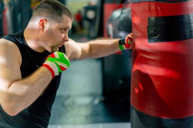 Bir boks kulübünde siyah tişörtlü yaşlı bir kas çalıştırıcı çok renkli bandajlarla boks yapıyor.