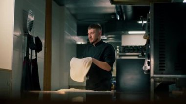 Profesyonel bir mutfakta, pizzacıda, siyah üniformalı genç bir şef havaya hamur krepi fırlatıyor.