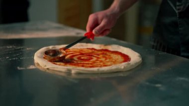 Bir pizzacıda profesyonel bir mutfakta siyah üniformalı genç bir şef pizzanın üzerine ketçap sürer.