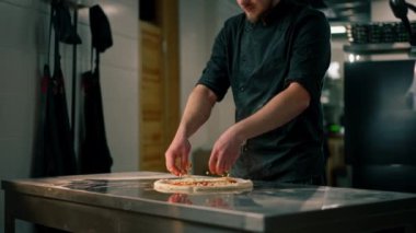 Profesyonel bir mutfakta bir pizzacıda siyah üniformalı genç bir şef pizzaya malzeme koyar.