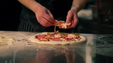 Bir pizzacının profesyonel mutfağında siyah üniformalı genç bir şef pizzanın içine malzeme koyar.