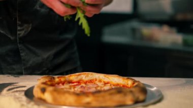 Bir pizzacının profesyonel mutfağında siyah üniformalı genç bir şef pizzanın içine malzeme koyar.