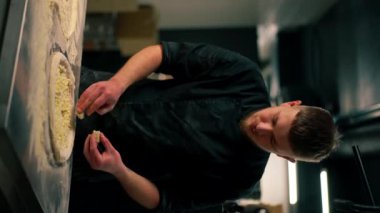 Pizzacıda profesyonel bir mutfakta dikey video. Siyah üniformalı genç bir şef pizzaya malzeme koyar.