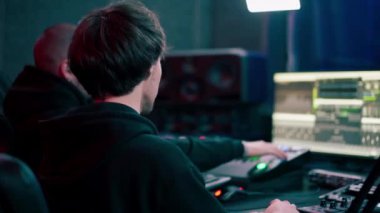 Kayıt odasının karanlığında, neon ışıklı bir stüdyo ile, bir ses kaydedicisi asistanına konsollar üzerinde nasıl çalışacağını öğretir.