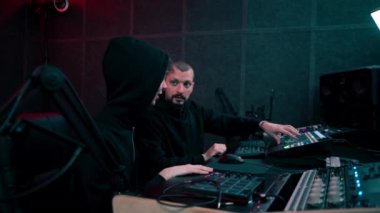 Kayıt odasının karanlığında, neon ışıklı bir stüdyo ile, bir ses kaydedicisi asistanına konsollar üzerinde nasıl çalışacağını öğretir.