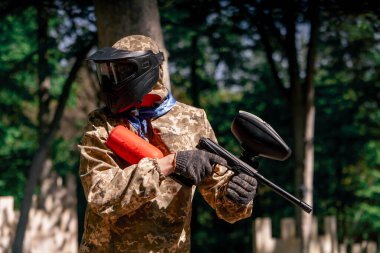 Vahşi doğada ağaçların yanında pintball üniformalı bir oyuncu elinde kırmızı balon takım elbisesi olan bir tabanca tutar.