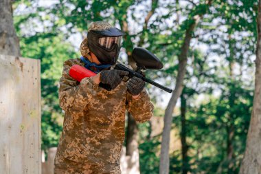 Vahşi doğada ağaçların yanında pintball üniformalı bir oyuncu elinde kırmızı balon takım elbisesi olan bir tabanca tutar.