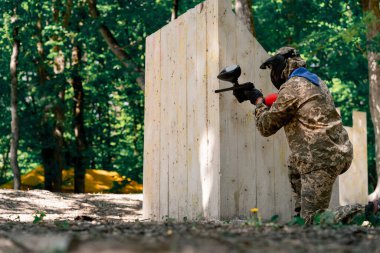 Vahşi doğada ağaçların yanında pintball üniformalı bir oyuncu elinde kırmızı balon takım elbisesi olan bir tabanca tutar.