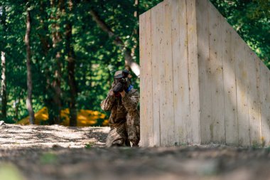 Vahşi doğada ağaçların yanında pintball üniformalı bir oyuncu elinde kırmızı balon takım elbisesi olan bir tabanca tutar.