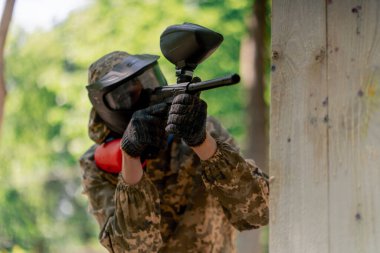 Vahşi doğada ağaçların yanında pintball üniformalı bir oyuncu kırmızı balon takım kıyafetleriyle bir tabanca tutuyor.