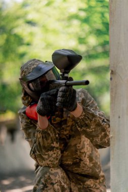 Vahşi doğada ağaçların yanında pintball üniformalı bir oyuncu kırmızı balon takım kıyafetleriyle bir tabanca tutuyor.