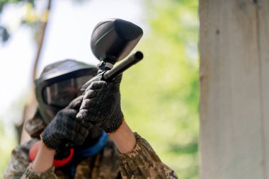 Vahşi doğada ağaçların yanında pintball üniformalı bir oyuncu kırmızı balon takım kıyafetleriyle bir tabanca tutuyor.