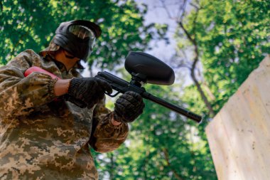 Vahşi doğada ağaçların yanında pintball üniformalı bir oyuncu elinde kırmızı balon takım elbisesi olan bir tabanca tutar.