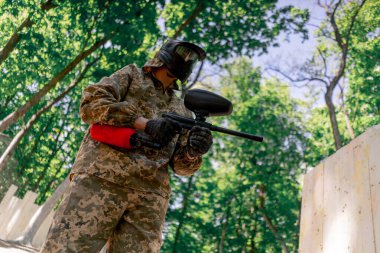 Vahşi doğada ağaçların yanında pintball üniformalı bir oyuncu elinde kırmızı balon takım elbisesi olan bir tabanca tutar.