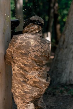 Vahşi doğada ağaçların yanında pintball üniformalı bir oyuncu kırmızı balon takım kıyafetleriyle bir tabanca tutuyor.