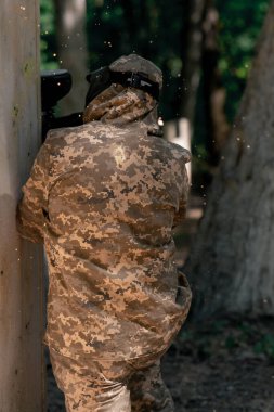 Vahşi doğada ağaçların yanında pintball üniformalı bir oyuncu kırmızı balon takım kıyafetleriyle bir tabanca tutuyor.