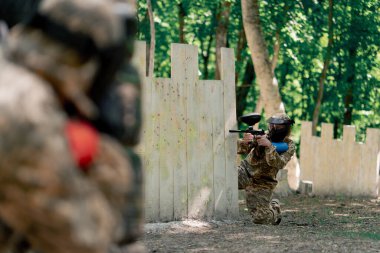 Vahşi doğada ağaçların yanında pintball üniformalı bir oyuncu mavi balon takım kıyafetleriyle bir tabanca tutuyor.