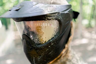 Formalı langırt oyuncusu yüzünde siyah koruyucu bir maskeyle bir paintball takımının yarışmasından kalma sarı bir lekeyle ormanın içinde.