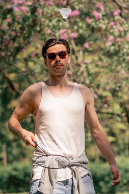 Dışarıda ağaçlarla dolu bir parkta kot pantolon ve beyaz tişörtlü yakışıklı bir esmer, bambinton oynamadan önce bir mekikle ısınan bir esmer.