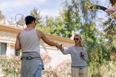 Dışarıda ağaçlarla kaplı bir parkta sarışın bir kızla genç bir esmer adam bambinton raketi eğitiyor en iyi arkadaşlar el sıkışıyorlar.