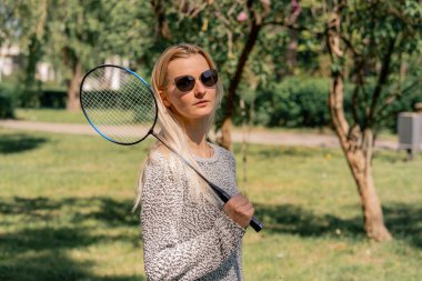 Dışarıda ağaçlarla kaplı bir parkta güneşli bir günde elinde bambinton raketi olan genç bir sarışın kız kameraya poz veriyor.