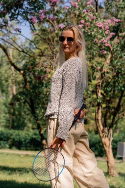 Dışarıda ağaçlarla kaplı bir parkta güneşli bir günde elinde bambinton raketi olan genç bir sarışın kız kameraya poz veriyor.