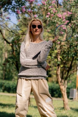 Dışarıda ağaçlarla kaplı bir parkta güneşli bir günde elinde bambinton raketi olan genç bir sarışın kız kameraya poz veriyor.