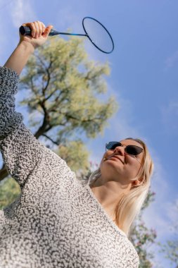 Ağaçlarla dolu bir parkta, küçük bir kız elinde bambinton bir raketle güneşli bir günde kameraya poz veriyor.