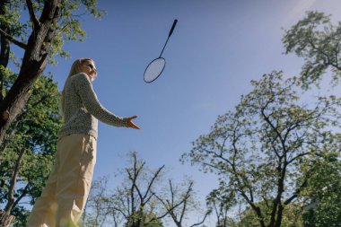 Güneşli bir günde, ağaçlarla kaplı bir parkta elinde bambinton raketi olan genç bir sarışın kız kameraya poz veriyor ve raketi havaya fırlatıyor.