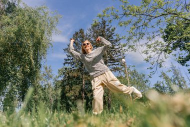 Teslim olma zeminine karşı ağaçlarla kaplı bir parkta genç sarışın bir kız raket hareketleriyle servis atıyor.
