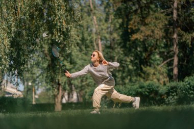 Teslim olma zeminine karşı ağaçlarla kaplı bir parkta genç sarışın bir kız raket hareketleriyle servis atıyor.