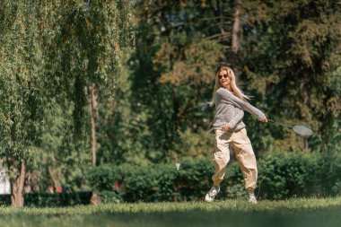 Teslim olma zeminine karşı ağaçlarla kaplı bir parkta genç sarışın bir kız raket hareketleriyle servis atıyor.