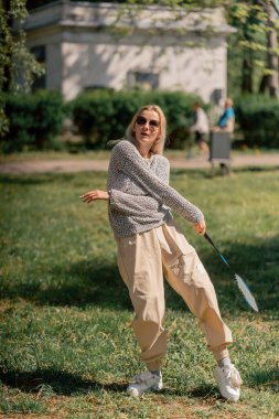 Teslim olma zeminine karşı ağaçlarla kaplı bir parkta genç sarışın bir kız raket hareketleriyle servis atıyor.