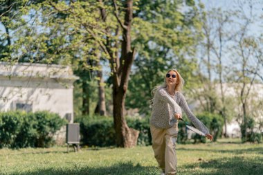 Teslim olma zeminine karşı ağaçlarla kaplı bir parkta genç sarışın bir kız raket hareketleriyle servis atıyor.