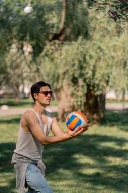 Dışarıda ağaçlarla dolu bir parkta beyaz tişörtlü ve güneş gözlüklü esmer bir adam voleybol topunu sokaktan önce ısıtıyor.
