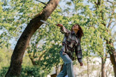 Dışarıda bir parkta ağaçların arasında teslim olma zeminine karşı esmer bir kız raket hareketleriyle servis atıyor.