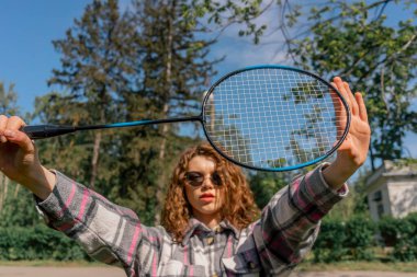 Parkta ağaçlarla çevrili bir yerde güneşli bir günde elinde bambinton raketiyle kameraya poz veren esmer bir kız var.