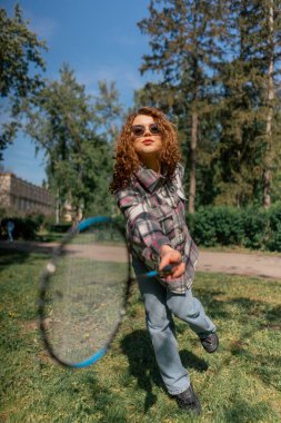 Dışarıda bir parkta ağaçların arasında teslim olma zeminine karşı esmer bir kız raket hareketleriyle servis atıyor.