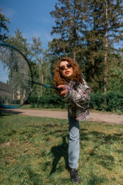 Dışarıda bir parkta ağaçların arasında teslim olma zeminine karşı esmer bir kız raket hareketleriyle servis atıyor.